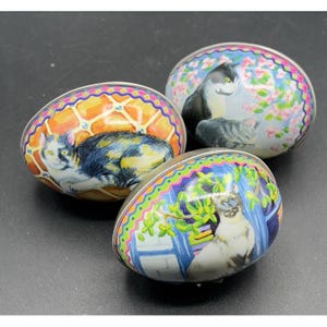 May include: Three decorative egg-shaped trinket boxes with cat illustrations. Each egg features a different cat design, set against colorful backgrounds with floral and geometric patterns. The eggs have silver rims.