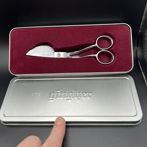 May include: A pair of silver-colored, curved-blade scissors rests inside a rectangular metal tin with a burgundy velvet lining. The tin is open, revealing the scissors and the embossed word "gingher" on the lid.