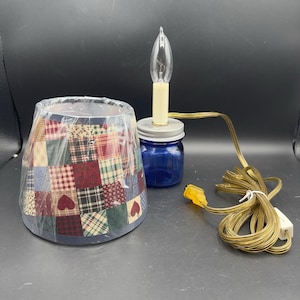 May include: A small lamp with a patchwork lampshade featuring red, green, and blue squares with heart accents. The lamp includes a blue jar base, a light bulb, and a cord with a plug.