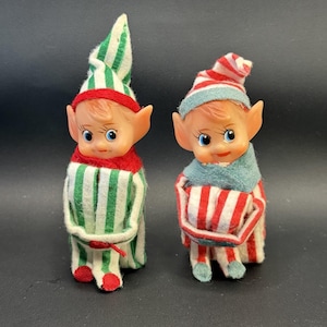 May include: Two small elf figurines with pointed ears and blue eyes. The left elf wears a green and white striped hat and outfit with red trim. The right elf wears a red and white striped outfit with a blue collar and a red and white striped hat.