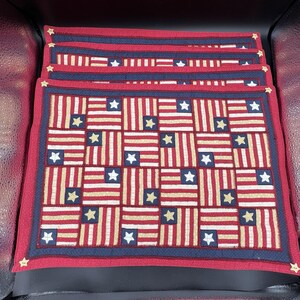 May include: Set of patriotic placemats featuring a patchwork design of the American flag. Each placemat has a red border, a navy blue inner border, and a repeating pattern of red and white stripes with gold stars. The placemats are square.