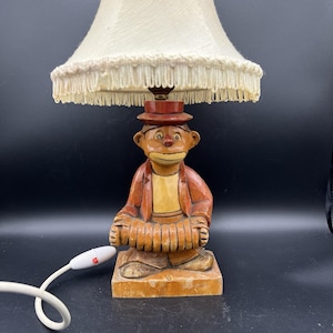 May include: A vintage wooden lamp featuring a carved figure of a monkey playing an accordion. The monkey wears a hat and jacket, with a cream-colored lampshade above. The lamp has a white cord with an on/off switch.