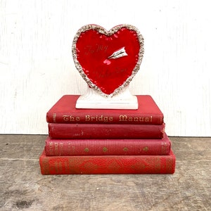 May include: A red heart-shaped ceramic figurine with a white base and a silver glitter trim. The heart has the words "To My Valentine" written on it with a silver arrow. The figurine is sitting on top of a stack of five red books. The top book has the title "The Bridge Manual" written on the spine.