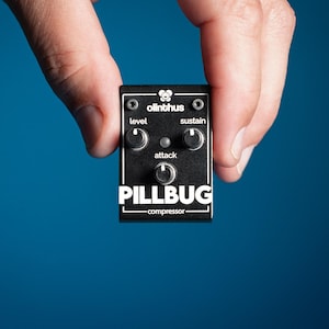 Puede incluir: Un pedal de compresor Pillbug negro con tres perillas etiquetadas como "level", "attack" y "sustain". El pedal tiene texto blanco y se sostiene sobre un fondo azul. La marca "olinthus" está en la parte superior.