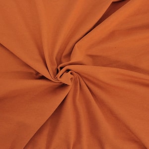 May include: A solid burnt orange fabric with a smooth, matte texture. The material is gathered in the center, creating soft, radiating folds and natural shadows that highlight the consistent, deep terracotta color across the entire surface.