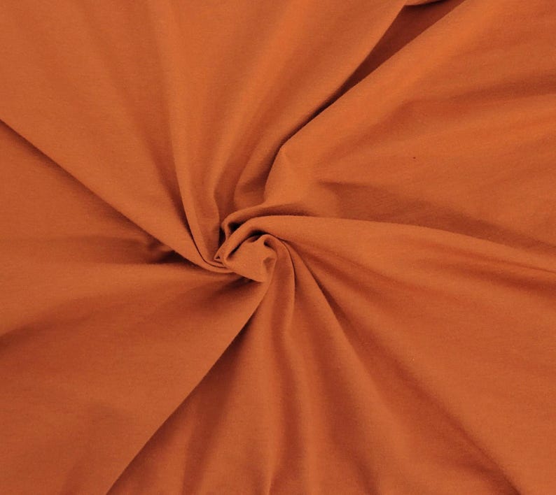 May include: A solid burnt orange fabric with a smooth, matte texture. The material is gathered in the center, creating soft, radiating folds and natural shadows that highlight the consistent, deep terracotta color across the entire surface.