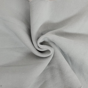 May include: A light gray fabric with a soft, matte texture, displayed in a series of deep, swirling folds and drapes. The material exhibits a consistent, neutral tone throughout, creating a series of shadows and highlights within the fabric's curves.