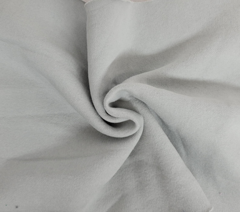 May include: A light gray fabric with a soft, matte texture, displayed in a series of deep, swirling folds and drapes. The material exhibits a consistent, neutral tone throughout, creating a series of shadows and highlights within the fabric's curves.