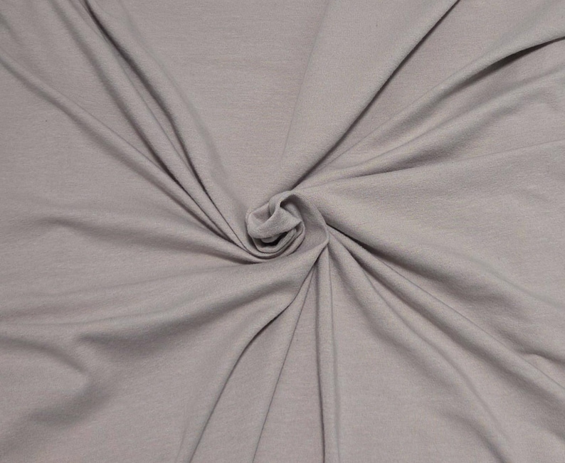 May include: A solid light gray fabric with a smooth, matte texture. The material is gathered in the center, creating soft, radiating folds and drapes that extend outward toward the edges, showcasing the consistent color and fine weave of the textile.