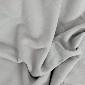 May include: A piece of light gray fabric with a soft, matte texture. The material is draped in loose, flowing folds, creating deep shadows and highlights across the surface. The edges of the textile appear raw and unfinished, showing the weave.