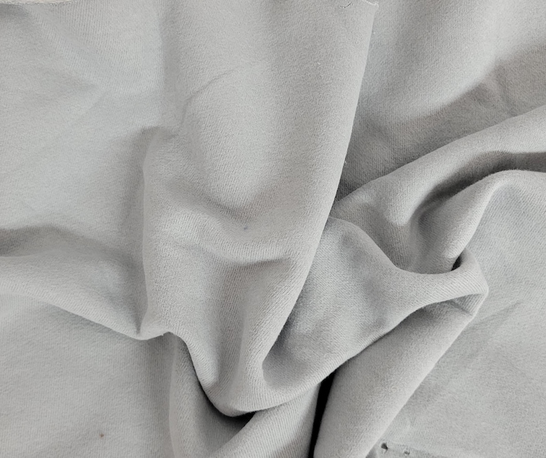 May include: A piece of light gray fabric with a soft, matte texture. The material is draped in loose, flowing folds, creating deep shadows and highlights across the surface. The edges of the textile appear raw and unfinished, showing the weave.