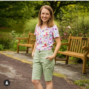 Pu&ograve; includere: Una donna indossa una camicia a maniche corte con stampa floreale e pantaloncini verde chiaro. &Egrave; in piedi davanti a due panchine di legno in un giardino.