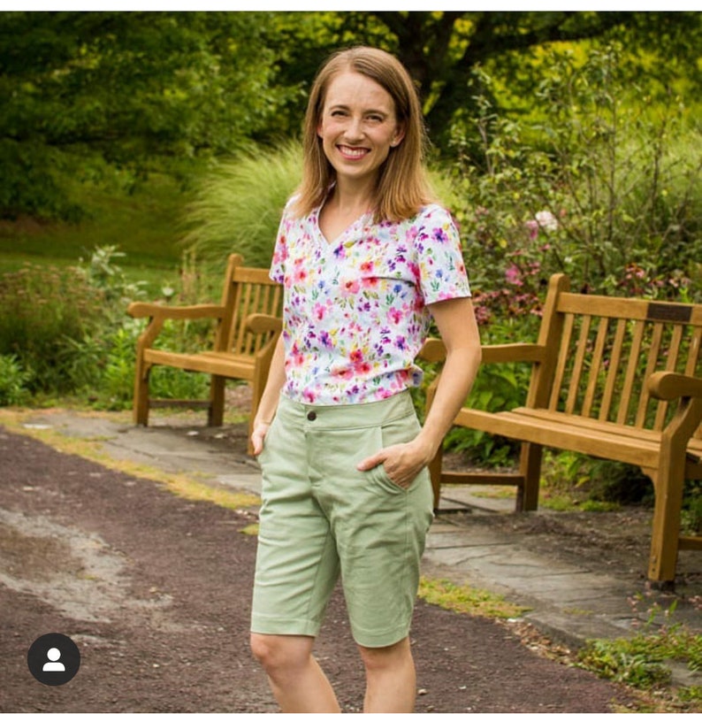 Pu&ograve; includere: Una donna indossa una camicia a maniche corte con stampa floreale e pantaloncini verde chiaro. &Egrave; in piedi davanti a due panchine di legno in un giardino.
