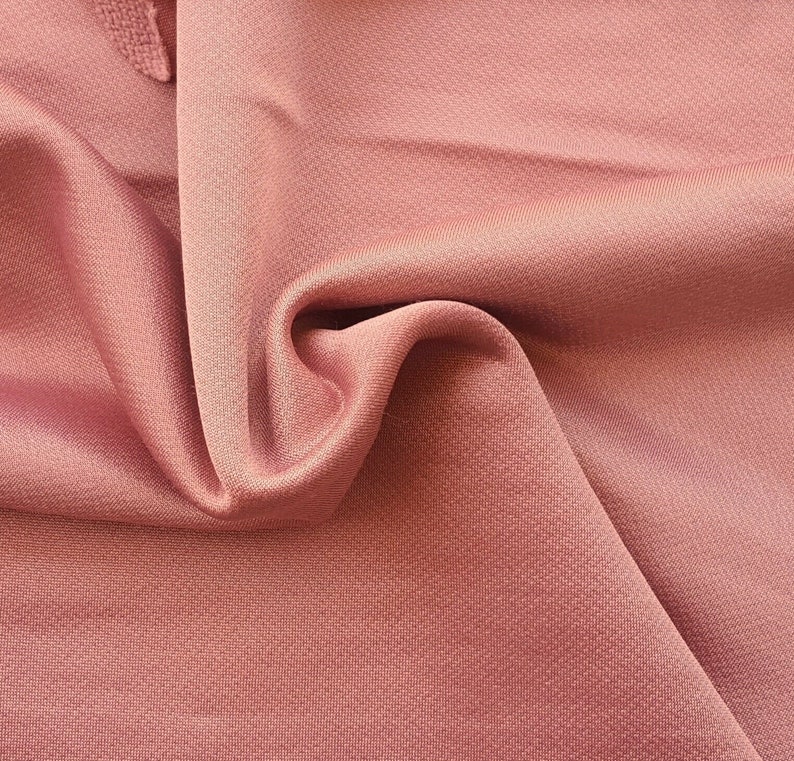 May include: Close-up of a soft, pink fabric with a subtle textured pattern. The fabric is draped and folded, showcasing its smooth and luxurious feel.