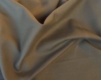 May include: A solid taupe-colored fabric with a smooth, matte finish and a tight weave. The material is draped in soft, flowing folds, creating natural shadows and highlights across the surface. The word koshtex is visible in the corner.