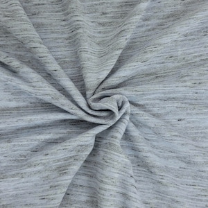 May include: Close-up of a light gray fabric with a subtle, textured appearance. The material has a heathered effect, with fine black and gray threads woven throughout. The fabric is gathered in a swirl, showing its drape and texture.