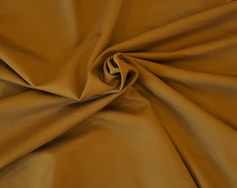 May include: Close-up of a golden-brown fabric with a soft, velvety texture. The material is gathered and swirled, creating a dynamic pattern of folds and shadows. The color is a rich, warm tone, reminiscent of autumn leaves.