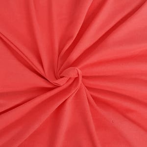 May include: A solid coral-colored fabric with a smooth, matte texture. The material is gathered in the center, creating deep, radiating folds and soft shadows that highlight the vibrant, uniform hue across the entire surface of the textile.