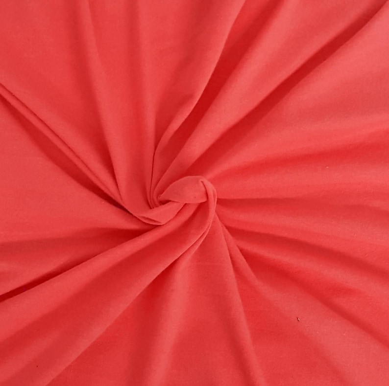 May include: A solid coral-colored fabric with a smooth, matte texture. The material is gathered in the center, creating deep, radiating folds and soft shadows that highlight the vibrant, uniform hue across the entire surface of the textile.