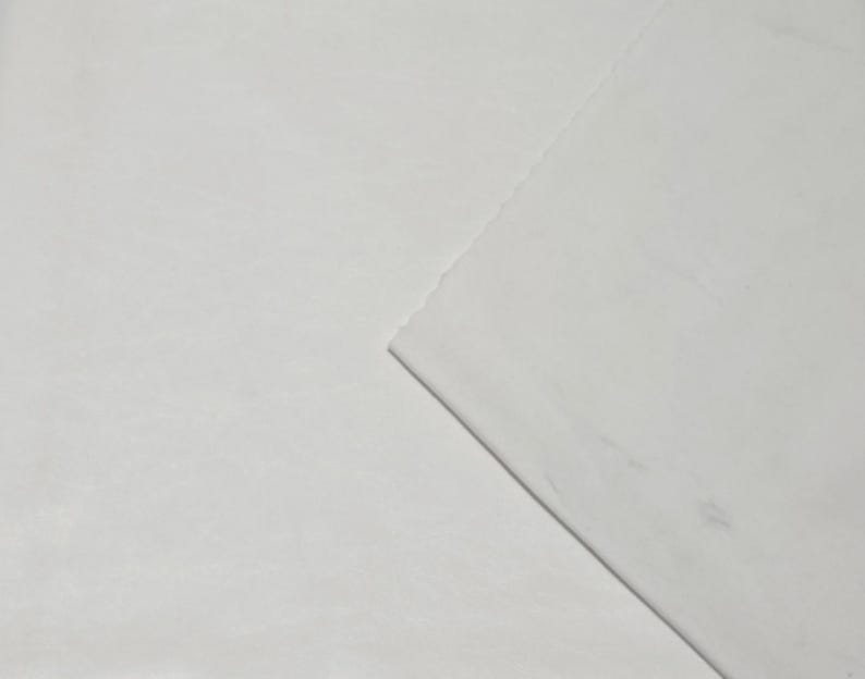 Pode incluir: Um close-up de tecido branco com uma superf&iacute;cie ligeiramente texturizada. O tecido &eacute; dobrado na diagonal, revelando uma borda em camadas. A ilumina&ccedil;&atilde;o cria sombras sutis, destacando a textura e as dobras.