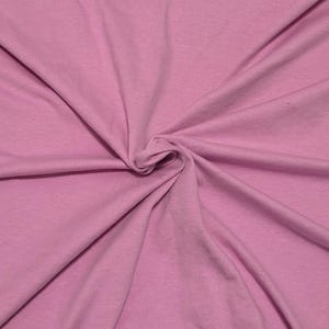 May include: A solid light pink fabric with a smooth, matte texture. The material is gathered in the center, creating soft, radiating folds and natural drapes that extend outward toward the edges of the frame, showcasing the consistent color.