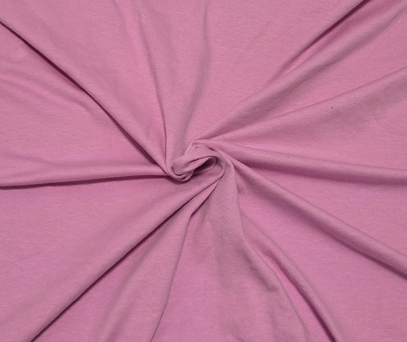 May include: A solid light pink fabric with a smooth, matte texture. The material is gathered in the center, creating soft, radiating folds and natural drapes that extend outward toward the edges of the frame, showcasing the consistent color.