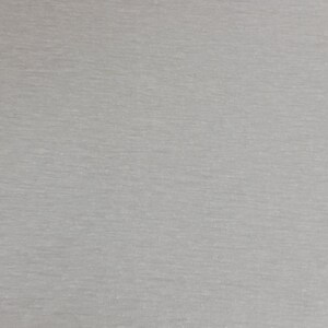 May include: A close-up shot of a light gray fabric with a subtle, textured appearance. The material has a slightly heathered effect, with variations in tone creating a soft, visual depth. The fabric appears to be a knit or jersey type.