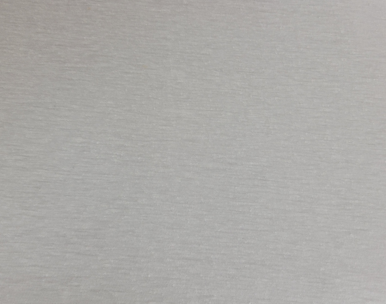 May include: A close-up shot of a light gray fabric with a subtle, textured appearance. The material has a slightly heathered effect, with variations in tone creating a soft, visual depth. The fabric appears to be a knit or jersey type.