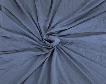 Może przedstawiać: Zbliżenie na niebiesko-szarą tkaninę. Materiał jest zebrany i skręcony w środku, tworząc efekt spirali. Tkanina wydaje się miękka i zwiewna, z widocznymi fałdami i zagnieceniami. Kolor to stonowany niebiesko-szary.