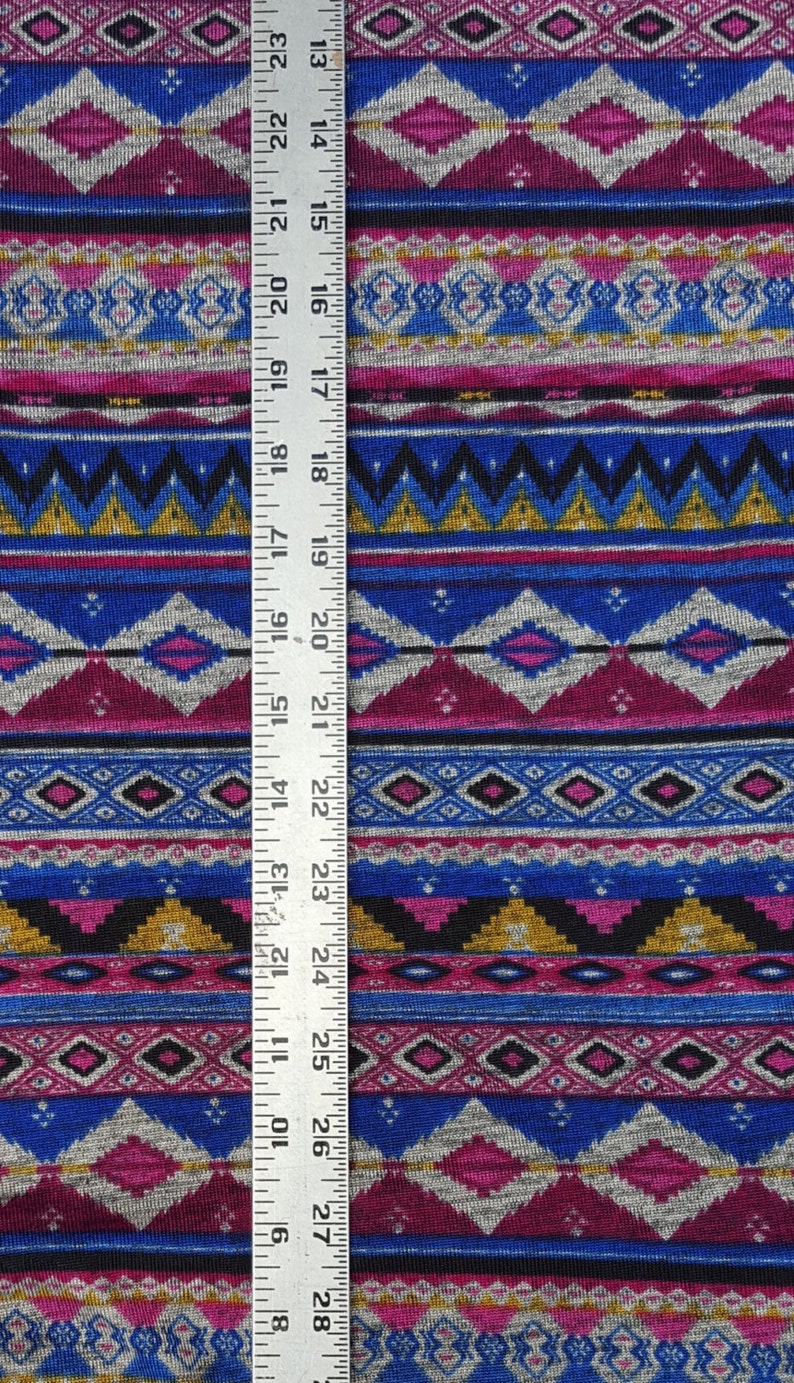 May include: A close-up of a colorful fabric with a geometric pattern. The fabric is a mix of pink, blue, gray, black, and gold. A ruler is placed next to the fabric to show the scale.
