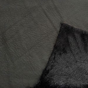 May include: Close-up of a dark gray fabric with a textured, wrinkled surface. A corner of the fabric is folded over, revealing a contrasting black, fuzzy underside. The material appears to be a faux fur or similar textile.