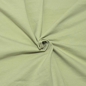 May include: A solid light sage green fabric with a smooth, matte texture. The material is gathered in the center, creating soft, radiating folds and natural drapes that extend outward across the entire surface of the textile material.