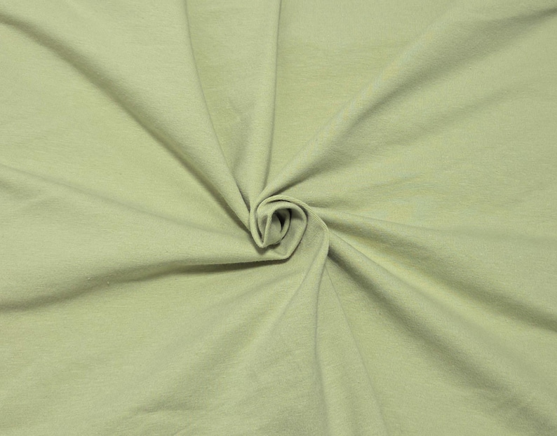 May include: A solid light sage green fabric with a smooth, matte texture. The material is gathered in the center, creating soft, radiating folds and natural drapes that extend outward across the entire surface of the textile material.