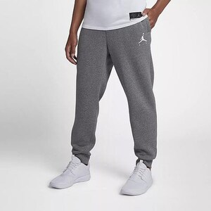 May include: Dark gray sweatpants with a white Jumpman logo on the left leg. The pants have a drawstring waist and cuffed ankles. The person is wearing a white t-shirt and white sneakers.