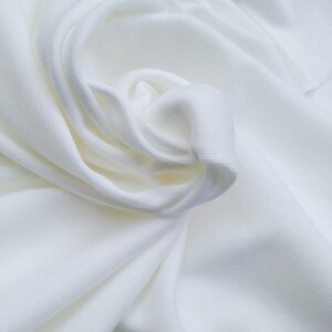 May include: Close-up of a white fabric with soft folds and a textured surface. The fabric appears to be a knit or woven material, with visible ribbing. The folds create shadows and highlights, adding depth to the image.