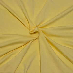 May include: A solid light yellow fabric with a smooth, matte texture. The material is gathered in the center, creating soft, radiating folds and natural drapes that extend outward toward the edges of the frame, highlighting the consistent color.