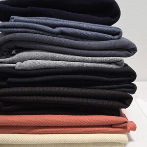 May include: A stack of folded polo shirts in various colors, including navy, blue, gray, black, coral, and cream. The shirts are neatly arranged, showcasing the different shades and textures of the fabric.