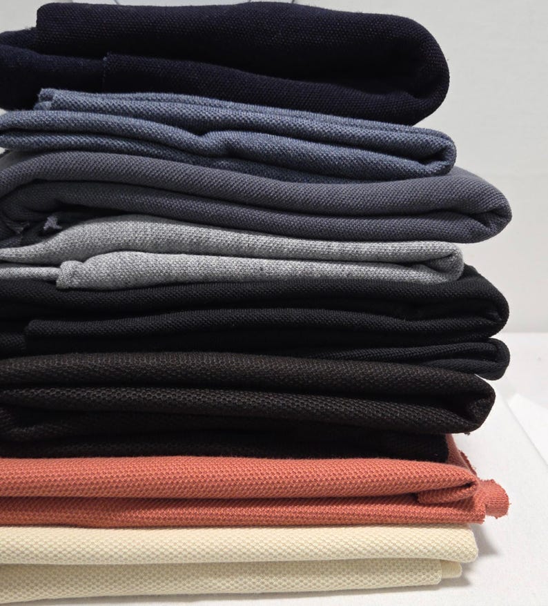 May include: A stack of folded polo shirts in various colors, including navy, blue, gray, black, coral, and cream. The shirts are neatly arranged, showcasing the different shades and textures of the fabric.