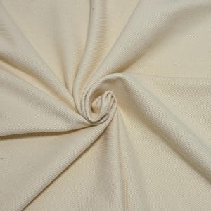 May include: Cream-colored fabric with a textured, woven pattern. The fabric is arranged in soft folds, with a central swirl. The material appears to be thick and durable, suitable for upholstery or clothing.