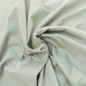 Puede incluir: Una tela de color verde salvia claro s&oacute;lido con una textura suave y mate. El material est&aacute; recogido en el centro, creando pliegues suaves y fluidos con sombras profundas que resaltan la ca&iacute;da y el color uniforme de la superficie textil.