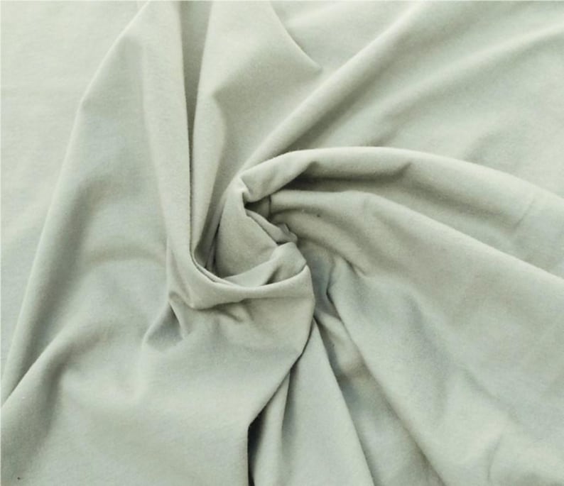 Puede incluir: Una tela de color verde salvia claro s&oacute;lido con una textura suave y mate. El material est&aacute; recogido en el centro, creando pliegues suaves y fluidos con sombras profundas que resaltan la ca&iacute;da y el color uniforme de la superficie textil.