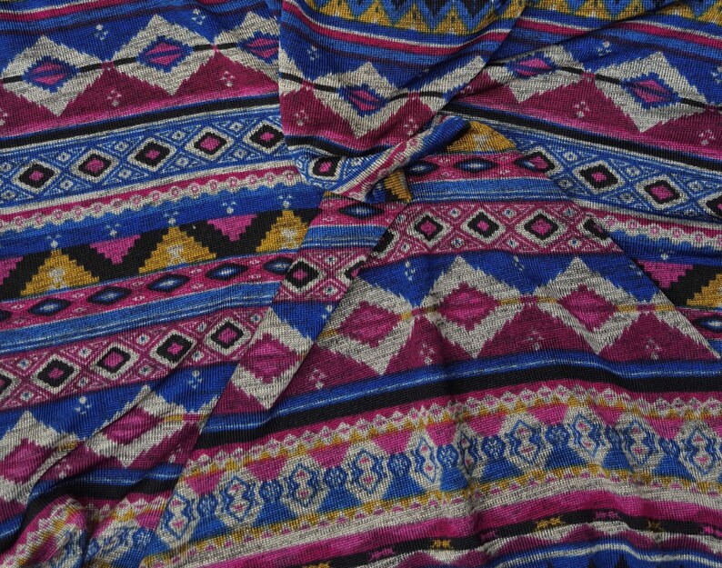 May include: A close-up of a fabric with a colorful geometric pattern. The pattern features shades of blue, pink, gold, and black. The fabric is woven with a textured surface.