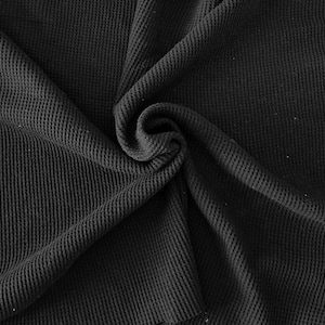 May include: Close-up of a black waffle knit fabric. The fabric is folded and draped, creating a textured surface with a spiral in the center. The material appears soft and has a subtle sheen, suggesting it could be used for clothing or home textiles.