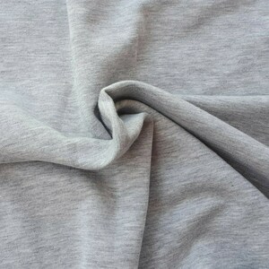 May include: Close-up of a light gray fabric with a soft, textured appearance. The fabric is draped and folded, creating shadows and highlighting the material's subtle variations in tone. The overall effect is one of comfort and gentle movement.