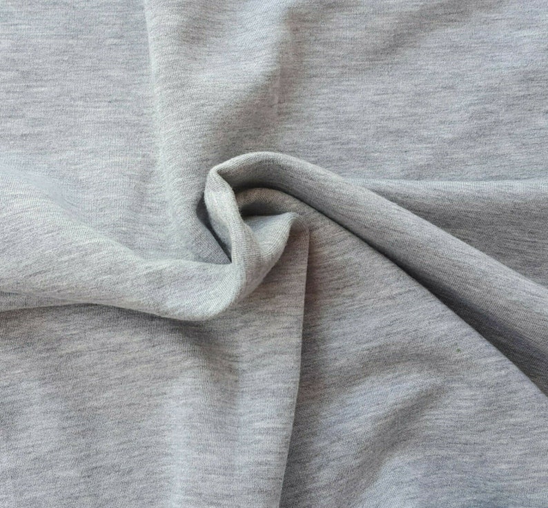 May include: Close-up of a light gray fabric with a soft, textured appearance. The fabric is draped and folded, creating shadows and highlighting the material's subtle variations in tone. The overall effect is one of comfort and gentle movement.