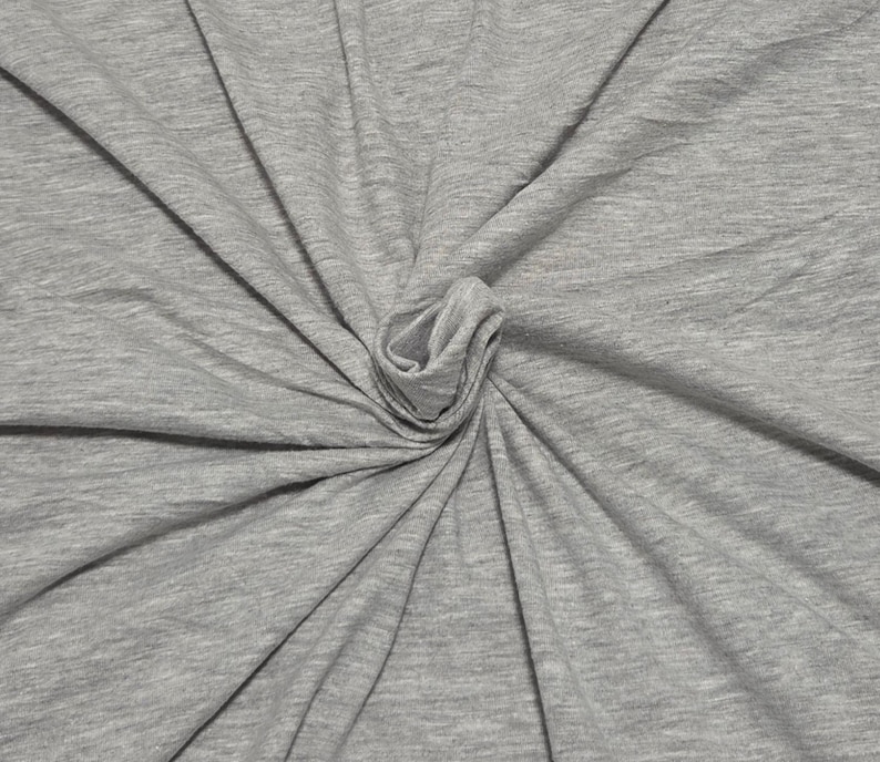May include: A heather gray fabric with a soft, jersey-like texture. The material is gathered in the center, creating radiating folds and drapes that extend outward toward the edges, showcasing the consistent light gray color and fine knit pattern.