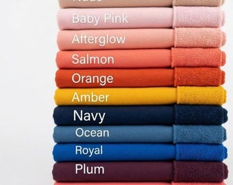 May include: A vertical stack of twelve folded rectangular towels in various colors, each labeled with white text: Nude, Baby Pink, Afterglow, Salmon, Orange, Amber, Navy, Ocean, Royal, Plum, and Coral. The towels feature a smooth texture on the main body and a textured, looped fabric border on the right side.