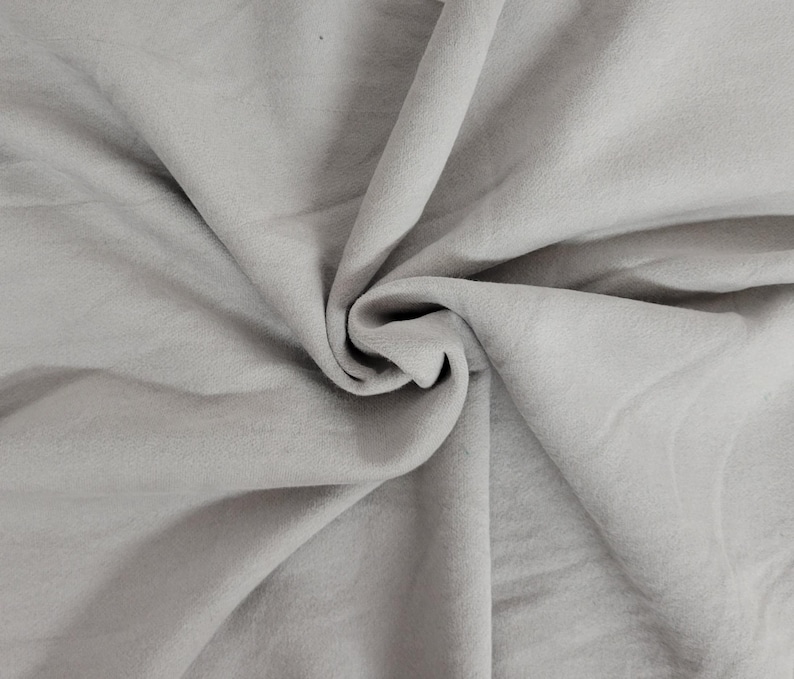 May include: A light gray fabric with a soft, matte texture, featuring deep, flowing folds and natural drapes that create a series of shadows and highlights across the surface, emphasizing the smooth, consistent color and pliable nature of the material.