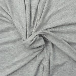 Puede incluir: Una tela jaspeada de color gris claro con una textura suave similar al punto. El material est&aacute; recogido en el centro, creando pliegues y drapeados naturales que resaltan las sutiles variaciones de las fibras grises claras y oscuras.
