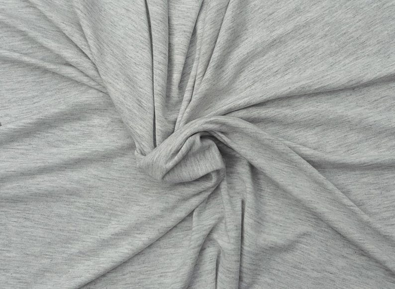Puede incluir: Una tela jaspeada de color gris claro con una textura suave similar al punto. El material est&aacute; recogido en el centro, creando pliegues y drapeados naturales que resaltan las sutiles variaciones de las fibras grises claras y oscuras.